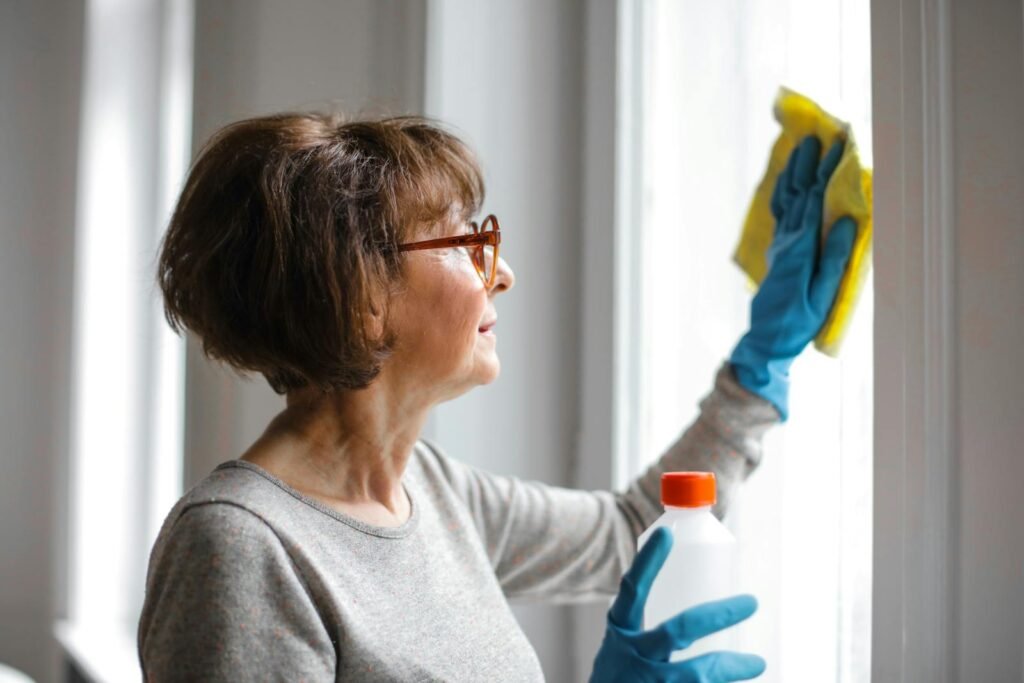 Limpieza Abella La Coruña Elderly woman cleansing window indoors using gloves and cleaner.